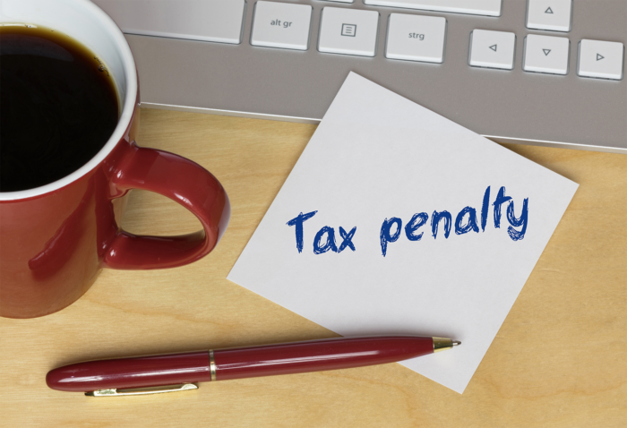 Desk with a cup of coffee, a red pen, the bottom of a keyboard, and a Post-It note that says 'tax penalty'